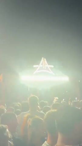 A nighttime outdoor event with a large crowd of people. A wooden structure with a neon sign displaying the letters "AA" is visible in the background. The crowd appears to be dancing and enjoying the atmosphere. Lighting effects and a bright vertical light source are present, contributing to a lively ambiance. The video is shaky, suggesting it was filmed by someone within the crowd.