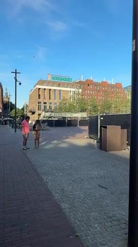 A vertical video shows an outdoor scene with two people standing in the foreground on a brick sidewalk, facing away from the camera towards a paved square. The person on the left is wearing a pink t-shirt and blue shorts. The person on the right is wearing a leopard print dress and carrying a bag. In the background, there is a multi-story brick building with the word "HOOGHOUDT" visible near the top. Several people are seated on a concrete ledge in front of the building, and there are black construction barriers and some metal structures in the middle ground. The sky is blue with a few clouds.
