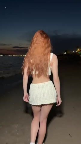 A young woman with long, wavy red hair is walking on a sandy beach at dusk. She is wearing a matching white knitted crop top and mini skirt. The sky is a gradient of dark blues and hints of orange from the setting sun. Behind her, the sea stretches out, and in the distance, lights suggest a populated area. She turns to face the camera, poses, then turns to her side.