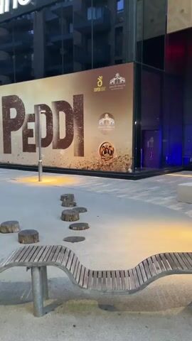 The video shows an outdoor urban area with modern architecture. In the background, a large banner displays the word "PEDDI" in large, distressed letters, with smaller circular images of what appear to be movie posters or promotional material on the right side of the banner. Above the banner, a portion of a building with the text "cineworl" and a red star logo is visible, suggesting it's a cinema. In the foreground, there are several wooden stumps arranged in a path, a curved wooden bench, and planters with small trees. Two people are walking away from the camera down a paved walkway lined with shops and residential buildings. The lighting suggests it is either dusk or early evening.