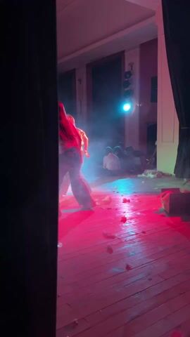 Here is a description of the video:

Two women dance on a wooden stage strewn with debris. The stage is bathed in alternating red, blue, and green light, with visible smoke effects. Some seated observers are visible at the back of the stage. The scene is framed by dark curtains on either side, providing a glimpse of the performance from the wings.