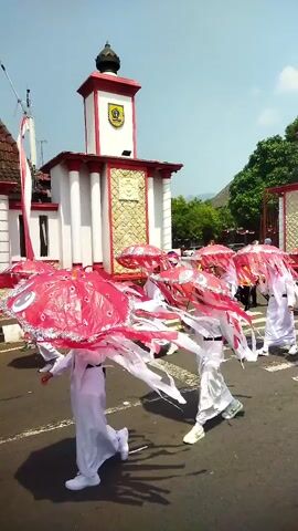 A group of people in white robes and red hats are marching down a street carrying large red umbrellas. They are participating in a parade, possibly a cultural event. The umbrellas are decorated with white patterns and fringe. The people are walking in unison, with some of them holding their arms out to the sides. The background shows a building with a red roof and white walls, and a flagpole with a red and white flag. The sky is blue and clear, and there are trees in the distance. The street is paved and there are some shadows on the ground.