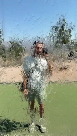 A person wearing a white t-shirt and shorts is standing behind a waterfall. The person is looking at the camera and waving. The background shows trees and a blue sky. The water droplets obscure some of the details of the person and the background.
