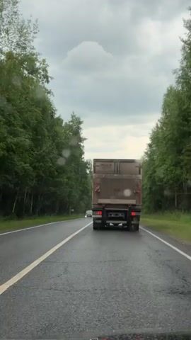 The video captures a point-of-view shot from inside a vehicle driving on a wet, paved road lined with dense green trees on both sides. The sky is overcast with gray clouds, suggesting a possibly rainy or post-rain condition.

The primary subjects are a large, boxy truck in front, moving in the same direction, and a smaller car approaching from the opposite direction. The truck is brown and appears to be a cargo truck, with its rear visible. The car, a dark sedan, has its headlights on and is clearly visible as it passes the camera vehicle. The road has a white solid line marking the center and a broken white line indicating a lane separation. There are visible water droplets on the windshield of the camera vehicle, further confirming wet weather.

The scene is set in a rural or semi-rural environment, suggested by the abundance of trees and the relatively empty road. The time of day appears to be daytime, but the overcast conditions obscure direct sunlight. There are no people visible. No text is legible in the video. The overall mood is one of transit and travel under less-than-ideal weather conditions.