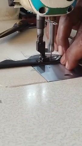 A close-up shot shows a person's hand guiding a dark piece of fabric under the needle of a white sewing machine. The needle is in motion, stitching the fabric. The sewing machine has a metal plate beneath the needle and a foot pedal visible in the background. The fabric appears to be a hem being sewn.