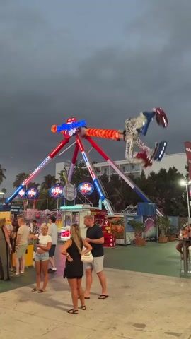 A tall, red, white, and blue amusement park ride, possibly a pendulum ride, is in motion. The ride has lights that flash in red, white, and blue patterns. The sky is cloudy and appears to be dusk. Below the ride, a group of people are standing and watching. One woman in the foreground wears a black dress and sandals, and a man next to her wears a white t-shirt and shorts. Further back, other people, including a woman in a white top and denim shorts, are visible. Several amusement park stalls and structures are present behind the people, with various signs and colorful displays. The ground appears to be paved.