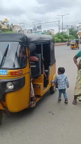 A young boy stands on the side of a road next to a yellow auto-rickshaw. A woman in a green sari is getting into the auto-rickshaw, while another woman in a red and white patterned outfit holds a phone and a black bag. Traffic is visible in the background, including other auto-rickshaws and cars. The sky is overcast.