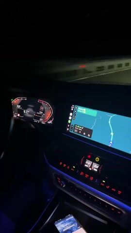 The video shows the interior of a car at night. The dashboard displays are illuminated, with the digital speedometer showing speeds between 126 and 115. The central infotainment screen shows a navigation map with a route highlighted and estimated arrival time. The ambient interior lighting is blue. A smartphone is visible in the center console. The car appears to be in motion on a road, as indicated by the blurred lights outside the car.