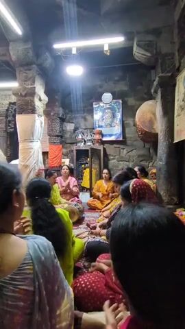 A group of women are gathered indoors, possibly in a temple, for what appears to be a religious ceremony or gathering. They are seated on the floor in a semi-circle, facing towards the front of the room. A framed picture of a deity is visible on the wall at the back, with some religious paraphernalia arranged below it. Some of the women are clapping their hands, while others are in prayer or listening attentively. The environment suggests a traditional setting with stone pillars and a low ceiling, illuminated by artificial lighting. The women are dressed in colorful traditional attire.