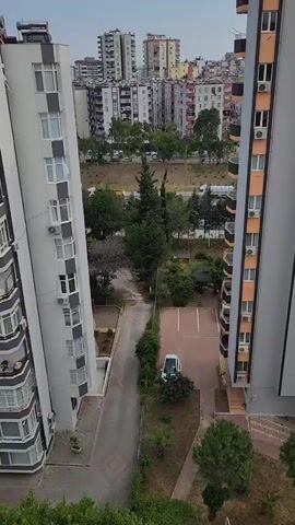 The video shows a view from a high vantage point looking down into an urban environment. The foreground features the sides of two tall apartment buildings, one on the left and one on the right, both with balconies and windows. Between the buildings, a narrow asphalt path curves downwards. To the right of the path, there is a small paved parking area with one white car parked and a small green gazebo. Further down, there is a patch of grass with some trees. Beyond this, there is a green area with more trees and a road with a few vehicles. In the background, a dense cluster of multi-story buildings and apartment blocks creates a city skyline under a cloudy sky. The overall scene depicts a residential area within a city.