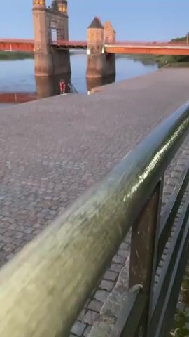 The video shows a wide cobblestone pathway bordering a river. In the background, a bridge with two stone towers spans the river. The bridge has an orange railing. A person in a red jacket stands near the water at the base of the bridge towers. The sky is a dusky blue, suggesting either dawn or dusk. The camera is focused on a metal railing in the foreground, which is slightly out of focus.