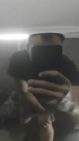 The video shows a person in a black t-shirt and shorts taking a selfie in a mirror. The person is crouching and holding a black smartphone. The background is blurry and appears to be an indoor environment. The mirror is reflecting other objects in the room, including what looks like a window with blinds and some shelves. The video transitions to a close-up of a black, glossy surface, possibly a car door, with newspaper and masking tape around the edges.