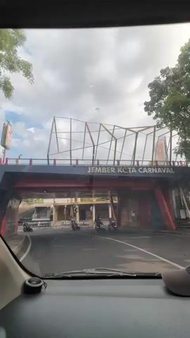 The video captures a view from inside a vehicle driving under an overhead structure. The structure appears to be a pedestrian bridge or overpass, painted in vibrant colors of red and yellow with a dark blue railing. Text on the blue railing reads "JEMBER KOTA CARNAVAL". The scene below the overpass shows a street with several motorcycles being ridden by people. Buildings line the street, some with signs, suggesting a commercial or residential area. The weather appears to be partly cloudy, with sunlight filtering through. The overall atmosphere is that of a typical day in a bustling Indonesian city.