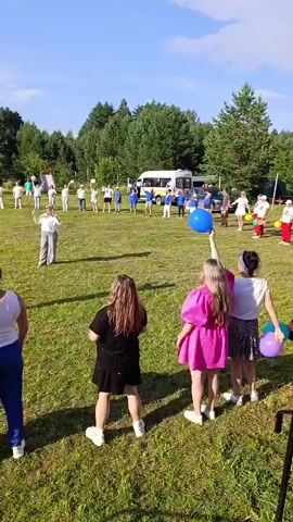 In an outdoor grassy area with trees and a stage in the background, a group of people are gathered on a sunny day. Many of them are holding balloons and raising them in the air. In the foreground, three women are facing away from the camera, each holding a balloon. The woman on the left holds a white balloon, the middle woman holds a white balloon, and the woman on the right holds a purple balloon. The women are wearing casual clothing. In the mid-ground, a larger group of people, including children and adults, are also holding balloons and appear to be participating in an activity. Some are wearing white shirts, while others wear blue. Two white vans are parked in the background. The overall atmosphere seems to be a festive outdoor event or gathering.