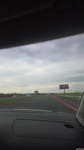 The video shows a first-person perspective from inside a car driving on a multi-lane highway. The car's dashboard is visible at the bottom of the frame, with the windshield framing the view ahead.

The road is a wide, paved highway with multiple lanes, and distinct red markings line the shoulders on both sides. Guardrails separate the road from the surrounding landscape. In the distance, other vehicles can be seen, but the highway appears relatively clear.

The sky is overcast with a blanket of grey clouds, suggesting either an early morning, late afternoon, or generally cloudy day. The landscape adjacent to the highway features green fields and some patches of bright yellow, possibly indicating blooming rapeseed or canola fields. Power lines and utility poles are visible stretching across the landscape in the distance.

Two billboards are visible alongside the highway. The one on the left shows the partial text "KAPITAL..." along with a person's face. The billboard on the right also features a person's face, possibly a celebrity or public figure, with text below that is difficult to decipher due to distance and resolution, but appears to be in Cyrillic script, consistent with the specified location of Krustina, Bulgaria.

The activity depicted is straightforward driving on a highway under cloudy conditions.