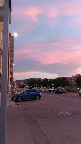 The video shows a street scene at dusk with a pastel sky. A blue SUV is parked on the left side of the road, and several other cars are driving by. Buildings and trees are visible in the background, and the sky is filled with pink and blue clouds. The video is shot from a low angle, looking up at the sky.