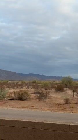 The video shows a wide shot of a desert landscape under a cloudy sky. The terrain is mostly flat with low-lying scrub brush and scattered trees. In the distance, there are mountains. A dirt road or path runs horizontally across the middle ground, with a low brick wall in the foreground. There are a few fence posts visible in the landscape. The lighting suggests it is either early morning or late evening, or possibly a heavily overcast day.