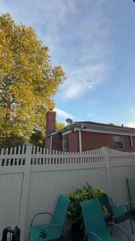 A video captures a satellite dish mounted on the roof of a brick house. The dish is gray and points upwards towards the blue sky, where thin white clouds are visible. To the left of the dish, a tall brick chimney rises above the house. A large tree with yellow-green leaves partially obstructs the view of the sky. A white picket fence runs horizontally across the lower portion of the frame, with greenery visible behind it in the foreground. Electrical wires are strung across the sky. The sun is shining, casting a warm light on the scene. The video is shot from a low angle, looking up at the house and sky.