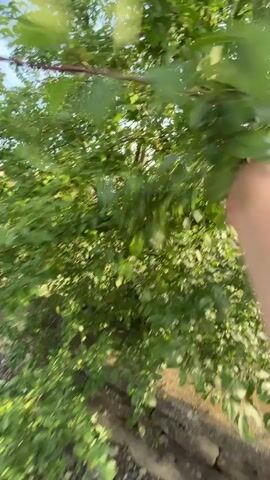 The video opens with a first-person view of a hand holding several small, round, green objects, possibly berries or seeds, with blurred railway tracks and green foliage in the background. The camera then pans up to show a young man wearing a light blue t-shirt with "ZY WKND" printed on the front. He is outdoors under a bright, sunny sky with visible power lines overhead.

The man looks upwards, smiling slightly, and then tilts his head back further, closing his eyes as if basking in the sun or observing something above him. In the background, a light-colored building with traditional architecture, several large green trees, and a green metal fence are visible. The ground is an unpaved mix of gravel and dirt. Another person is partially visible in the background. The bright sunshine suggests it is daytime, possibly late afternoon, in a rural or semi-rural setting, consistent with the provided location context of Lozarevo, Bulgaria.