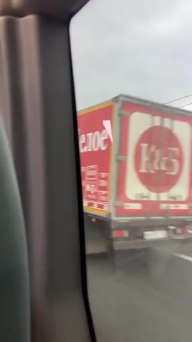 From the interior of a moving vehicle, a fleeting view through the side window reveals a red truck with white Cyrillic text, identified as a truck for the Russian retail chain "Krasnoe & Beloe" (Red & White). The scene quickly transitions to a highway landscape featuring a gray asphalt road, a metal guardrail, flat grassy fields, distant power lines, and a dense line of trees along the horizon under an overcast sky.