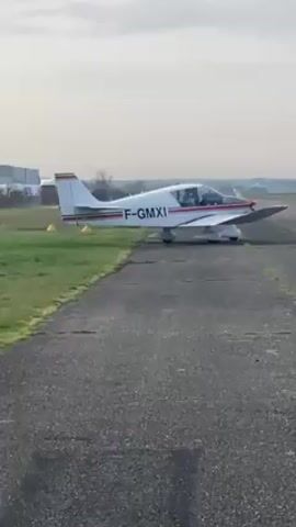 A small, white aircraft with red and black stripes taxis along a gray, weathered tarmac runway. The plane has the registration number F-GMXI on its fuselage. The propeller is spinning, indicating the engine is running. The runway is bordered by green grass on both sides. The sky is overcast and hazy, with buildings visible in the distant background. The camera is stationary and faces the plane directly as it moves away.