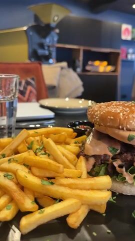 The image displays a close-up shot of a prepared meal, likely in a restaurant or café setting, located in Meisenheim, Germany.

The central subject is a substantial burger and a generous side of french fries, presented on a dark, possibly slate or ceramic, plate. The burger features a toasted sesame seed bun, a thick, irregularly shaped, dark brown patty, and fresh green lettuce. A creamy, pinkish-orange sauce is generously drizzled over the patty and lettuce. A wooden skewer holds the burger ingredients together. Behind the burger, a small, dark, round bowl is partially visible, likely containing an additional condiment or dipping sauce.

To the left of the burger, a pile of golden-brown french fries is seasoned with finely chopped green herbs, possibly parsley, adding a fresh visual element. A clear drinking glass, filled with water, is partially visible on the far left.

The background shows elements of the dining establishment. A table with a dark surface holds the meal. Further back, a decorative pillow with a geometric pattern in red, black, and white is visible on a chair or bench. Most notably, a large, industrial-looking coffee roaster, predominantly yellowish-gold and black, stands in the mid-background, suggesting that the establishment may be a café or roast its own coffee. Dark shelving units and other blurred interior details are visible further back, contributing to the ambiance of an indoor dining area.

The lighting suggests daytime or a well-lit interior, providing clear visibility of the food. No people are visible in the frame, and there is no discernible text. The overall scene captures an appetizing meal ready for consumption within a modern, casual eatery.