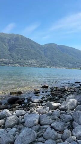This media file captures a serene lakeside scene with a prominent mountainous backdrop, likely in Locarno, Switzerland. The foreground is dominated by a rocky shore, with numerous smooth, grey stones of varying sizes scattered across the frame. The clear, calm water of the lake gently laps at the shore, with small ripples and reflections visible.

In the mid-ground, the lake stretches out, its surface shimmering under the sunlight. Across the water, the opposite shore is lined with a green, tree-covered mountainside, dotted with what appear to be buildings, suggesting a town or village nestled against the slopes. The mountains rise majestically, their peaks reaching towards the sky.

The sky above is a clear, vibrant blue, with a few wispy white clouds drifting lazily across it. The overall impression is one of tranquility and natural beauty. The lighting suggests it is daytime, likely late morning or early afternoon, given the bright sunlight and clear skies. There is no visible text in the media file. The scene evokes a sense of peace and offers a glimpse into a picturesque natural landscape.