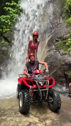A man and a woman pose for a photo on a red ATV in front of a waterfall. The man is in the driver's seat, wearing a black tank top, a red helmet, and smiling. The woman is standing behind him on the ATV, also wearing a red helmet, a pink top, patterned shorts, and has her arms raised in the air. The ATV is on a rocky surface with some water flowing around it. Behind them, a waterfall cascades down a rock face covered with lush green vegetation. The scene is lit by natural daylight, casting soft shadows on the surrounding greenery and creating a vibrant, tropical atmosphere.