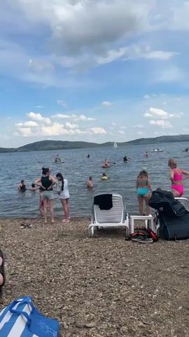 The video shows a crowded lakefront with many people swimming and participating in water activities. In the foreground, there is a rocky beach with several lounge chairs, bags, and a cooler. Several people are standing at the edge of the water, some talking and others wading in. In the water, there are numerous individuals and groups engaged in various activities, including swimming, paddleboarding, and floating on inflatable devices. A sailboat can be seen in the distance, along with other boats and watercraft. The sky is partly cloudy with the sun shining through, indicating a pleasant day. The overall scene depicts a lively summer recreational setting.
