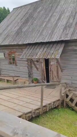 The video shows an old wooden mill building with a water wheel. The mill is constructed of rough-hewn logs and has a wooden roof. There is a wooden walkway leading to the entrance, and the water wheel is positioned to the side of the building. The surrounding environment is green and appears to be a park or rural setting with trees and grass. In the background, there are some people walking around and other wooden structures visible. The overall impression is of a historical or rustic site.