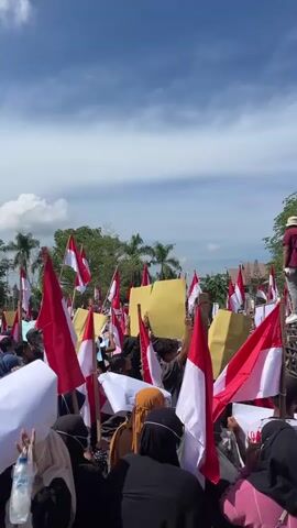 A vertical, medium shot captures a crowded outdoor gathering under a partly cloudy sky. The dominant colors are red and white, from numerous Indonesian flags being held aloft by the crowd. Many individuals are wearing headscarves, with some dressed in dark clothing and others in brighter colors like pink. In the background, there's a truck with people standing on top, some holding flags and what appear to be yellow signs. One person on the truck is holding a microphone. The atmosphere suggests a protest or rally. The camera is positioned within the crowd, looking forward towards the truck and the sky.