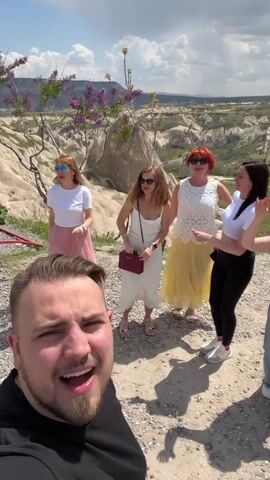 A group of approximately seven people, consisting of men and women, are captured in a selfie video cheering enthusiastically against a backdrop of unique geological formations in Göreme, Turkey. The man in the foreground is filming, his mouth wide open in an excited shout. The other group members behind him are also cheering, with their arms raised and mouths open, displaying expressions of joy and excitement. One woman in a red t-shirt has the partial text "AN" visible on it. The weather is overcast with clouds, but the scene is brightly lit. The landscape features light-colored, sculpted rock formations and some sparse vegetation under a vast sky. The overall atmosphere is lively and celebratory.