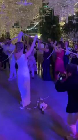 The video shows a group of people at a wedding. A woman in a white dress is standing in the center, holding ribbons in her hands with other women. There are several people around them, some holding up their hands with ribbons. In the background, there are men and women watching, some are filming. A photographer is visible in the foreground, taking pictures.  The setting is a dance floor with what appears to be light fixtures above. A white bouquet is on the floor near the bride.