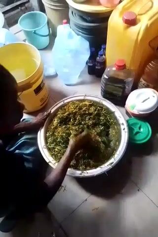 A person wearing black clothing is seated and mixing a large bowl of what appears to be leafy green vegetables with their hands. Another person, partially visible, adds a liquid from a plastic bottle with a red cap to the mixture. The setting appears to be a kitchen or storage area with various plastic buckets, jugs, bottles, and containers scattered around on the floor and stacked up. The lighting is dim, and the floor is tiled.