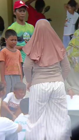 The video shows a group of children and adults gathered outdoors on a blue tarp. A woman in a light brown hijab and striped pants is facing away from the camera, with her hands on her hips. To her left, a young boy in an orange shirt is looking down. To the left of the woman, several children are sitting on the tarp. One girl in a white shirt and jeans is looking at the woman. Another child in a white shirt with "RONALDO" visible is holding a white balloon. A woman in a green and white patterned hijab is standing and holding a clipboard. Other people are visible in the background. The scene appears to be some kind of outdoor event or activity for children.