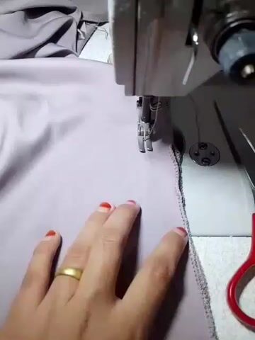 A close-up shot shows a person's hands operating a sewing machine. The sewing machine is in the upper right portion of the frame. A piece of purple fabric is being sewn, with the needle of the sewing machine piercing the fabric. The edge of the fabric has been serged. A pair of red-handled scissors is visible on the right side of the frame. The person's left hand is guiding the fabric, with a gold ring on their ring finger. The person's fingernails are painted red. The background is a white surface, likely a sewing table. The video is shot from a first-person perspective.