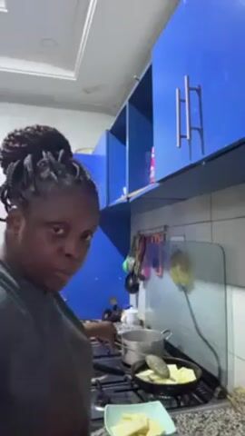 A woman is in a kitchen frying what appears to be plantains. She is standing in front of a gas stove with a frying pan on one burner containing plantain pieces frying in oil. A smaller pot with a strainer-like spoon rests on another burner. She uses a spatula to flip the plantains in the pan. The kitchen has blue cabinets and white tiled walls. Cooking utensils hang on a rack near the stove. The woman has her hair up in a bun and is wearing a casual shirt.