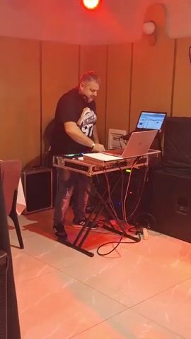 A DJ is standing at a table with his equipment, including laptops, mixing board, and headphones. He's wearing a black t-shirt, blue jeans, and sneakers. The room has pale yellow walls, and the floor is tiled in a pattern of pale yellow and white. There are speakers and other equipment around the DJ, as well as a chair on the left. The DJ is smiling and adjusting his equipment. There are multi-colored lights flashing above him.