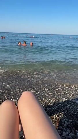 This is a first-person perspective video from a beach. The legs of the videographer are in the foreground, and they are sitting on a pebble beach. The sea is in the middle ground, with gentle waves lapping at the shore. There are several people in the water, swimming and playing. In the background, there is a pier with a building and umbrellas, and the sky is a clear blue. The sun appears to be shining brightly.