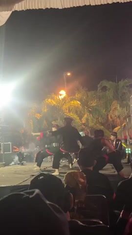 The video shows a nighttime outdoor performance on a stage. Several performers, dressed in black outfits with red accents and head coverings, are engaged in a synchronized martial arts or dance routine. Some performers hold wooden sticks. Smoke or fog is visible on the stage, illuminated by bright stage lights. In the background, palm trees are visible against a dark night sky, and a street light can be seen. The foreground shows the backs of an audience seated in chairs, observing the performance. The overall atmosphere appears to be festive or ceremonial.