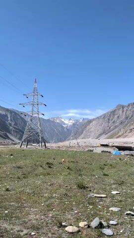 The video captures a wide shot of a mountainous landscape under a clear blue sky. In the foreground, a grassy area with scattered rocks transitions into a rocky, barren terrain. Several large, metallic electricity pylons with wires stretching across the frame dominate the midground. Behind the pylons, steep, gray mountains rise, with snow-capped peaks visible in the distance. The overall scene suggests a remote, high-altitude environment, likely a valley between mountain ranges, where power transmission infrastructure is present.