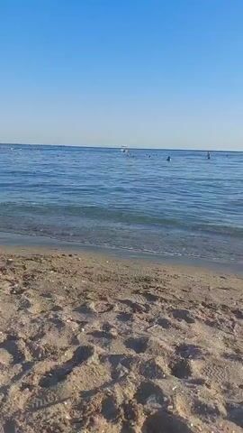 The video captures a beach scene on a clear, sunny day. The foreground is dominated by sand with footprints and some debris. The middle ground shows gentle waves lapping onto the shore. In the distance, the blue sea stretches to the horizon, meeting a clear blue sky. Several people are visible in the water, swimming or wading. Two boats are also present in the sea. The overall impression is a peaceful, recreational beach environment.