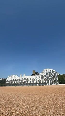 The video shows a long, white, modern building with multiple floors and balconies situated on a beach made up of small brown pebbles.  The sky is a clear, bright blue.  The building has a symmetrical design, with a central section that is lower and flanked by taller, curved sections. There are a few people visible on the balconies and walking along the base of the building.  Behind the building, some greenery can be seen, along with a glimpse of another building further up the hill.  The camera angle is low, emphasizing the beach and the size of the building.