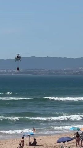 A helicopter flies over a beach, carrying a red bucket. The helicopter is white with red markings. The bucket is hanging below the helicopter by a cable. The beach is crowded with people, some of whom are lying on the sand and others are sitting under umbrellas. The ocean is blue and there are waves breaking on the shore. In the background, there is a city skyline and mountains. The sky is clear and blue. The video appears to have been shot on a sunny day.