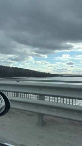A daytime video captured from a moving vehicle shows a scenic view of a wide body of water, likely a river or large lake, with a dark forest lining the distant bank. The sky is partly cloudy, featuring both large grey clouds and patches of bright blue sky with smaller white clouds. A metal guardrail and a portion of a car's side mirror are visible in the foreground, suggesting the vehicle is traveling on a bridge or elevated road alongside the water. The water appears calm, reflecting the light. The overall scene depicts a tranquil natural landscape under a dynamic sky. The location context indicates this is near Пальники, Russia.