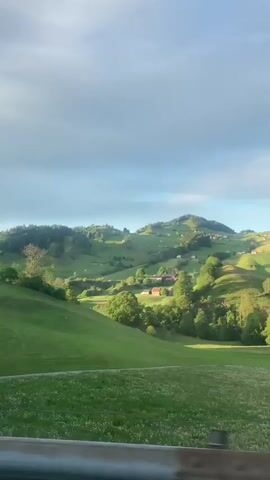 The video shows a scenic landscape of rolling green hills with scattered trees and small farmhouses, set against a sky with a mix of blue and fluffy white clouds. The camera appears to be moving, likely from a vehicle driving along a road, as suggested by the guardrail visible at the bottom. The footage is taken during a time of day when the lighting creates shadows on the hills.