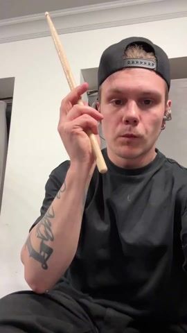 The video shows a man with tattoos on his left forearm, wearing a black t-shirt and a backwards black baseball cap. He is sitting and holding a drumstick, which he twirls and flips between his fingers. He makes eye contact with the camera throughout, making various expressions, including a smirk. The background consists of a white wall, a partially visible window with a curtain, and a gray rectangular panel. He also appears to be wearing in-ear headphones or earbuds.