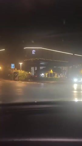 The video captures a nighttime drive from the perspective of inside a car, looking out through the windshield. The vehicle navigates a paved road lined with yellow and white curb markings, approaching a large, well-lit facility.

At the beginning, a building with an illuminated, decorative roofline featuring a stylized "E" logo is visible. To the left, a blue square sign with a white "P" indicates parking. Round, glowing streetlights illuminate the area, alongside some blurred greenery.

As the car proceeds, it turns towards a grand entrance. A prominent, brightly lit portico or canopy of a large building comes into view. The building's facade clearly displays the text "NVENTION CENTER" (the initial "CO" is out of frame). The entrance area beneath the canopy is brightly lit with numerous recessed lights. The scene is dark overall, with artificial lights providing the main illumination. No people are visible. The setting appears to be a modern convention center or a similar large venue, consistent with the location context of Agios Epiktitos, Northern Cyprus.
