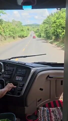 The video begins by showing the interior of a bus, with a hand on the gear shift. The camera pans forward through the windshield, revealing a two-lane road stretching into the distance. The road is lined with lush green trees and vegetation on both sides. In the distance, there are two cars, one red and one silver, and a third vehicle, possibly a van, further ahead. The sky is partly cloudy and the overall scene suggests a scenic drive through a rural or forested area. The camera remains focused on the road ahead for the duration of the clip.