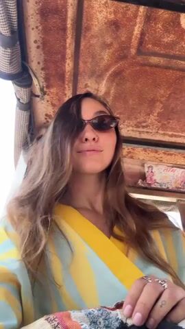 A young woman with long brown hair, wearing sunglasses and a yellow and blue shirt, is riding in an open-air vehicle, possibly an auto rickshaw. She is smiling at the camera and her hair is blowing in the wind. The interior of the vehicle has a rusty metal roof and patterned coverings. The background noises suggest the vehicle is moving on a street.